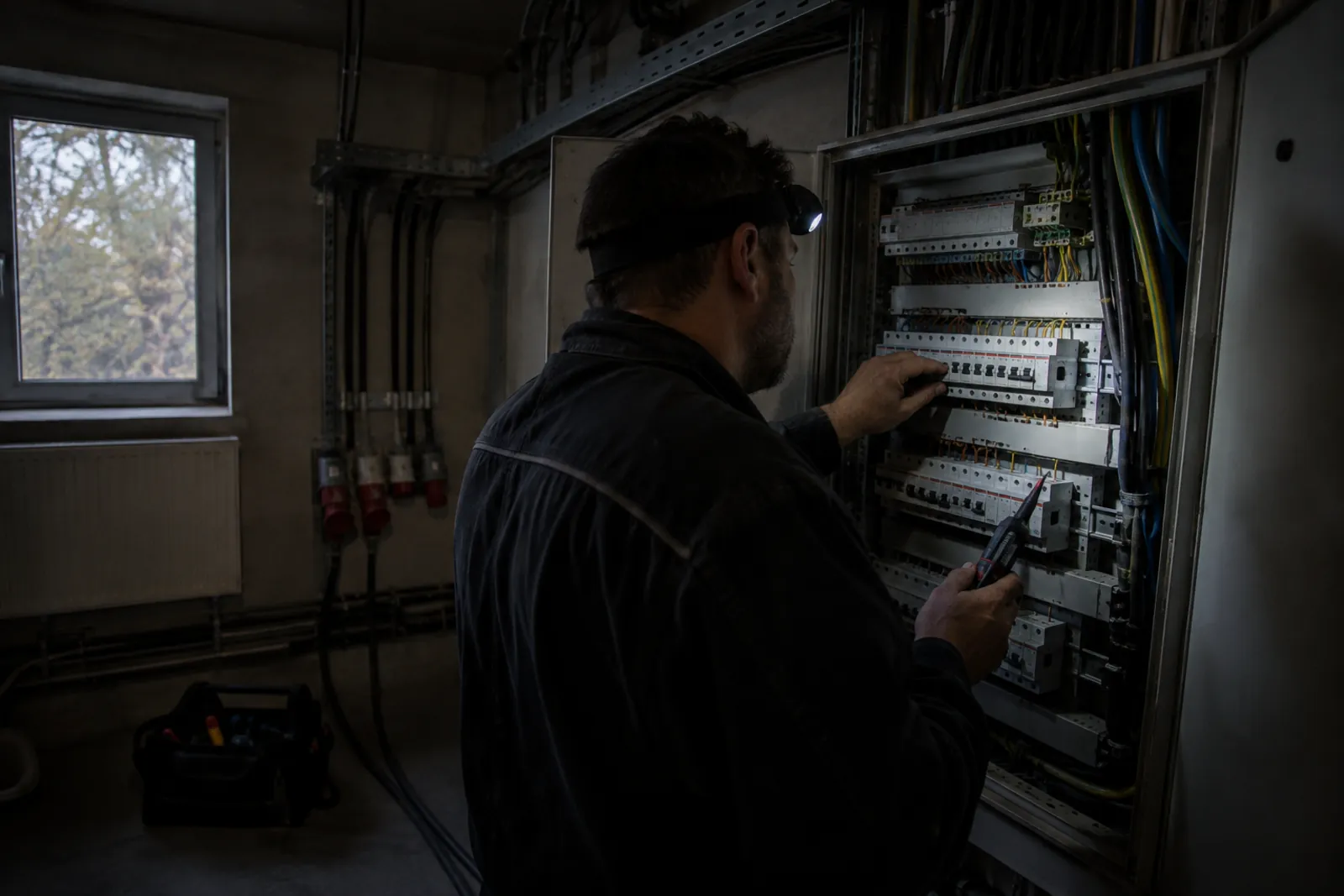 Elektrikář s čelovkou kontroluje jističe v otevřeném hlavním rozvaděči firmy, místnost je bez proudu